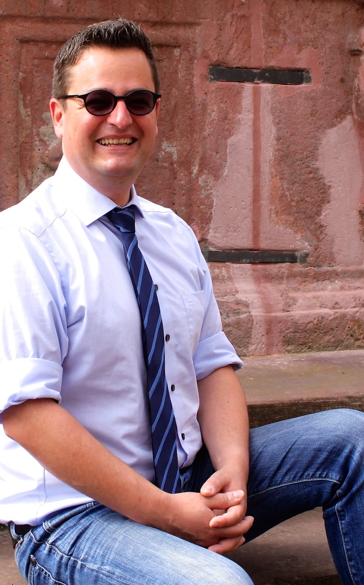 Michael Bailer Kirchenmusiker Michael Bailer sitzt im Freien mit weißem Hemd, Krawatte und Sonnebrille.